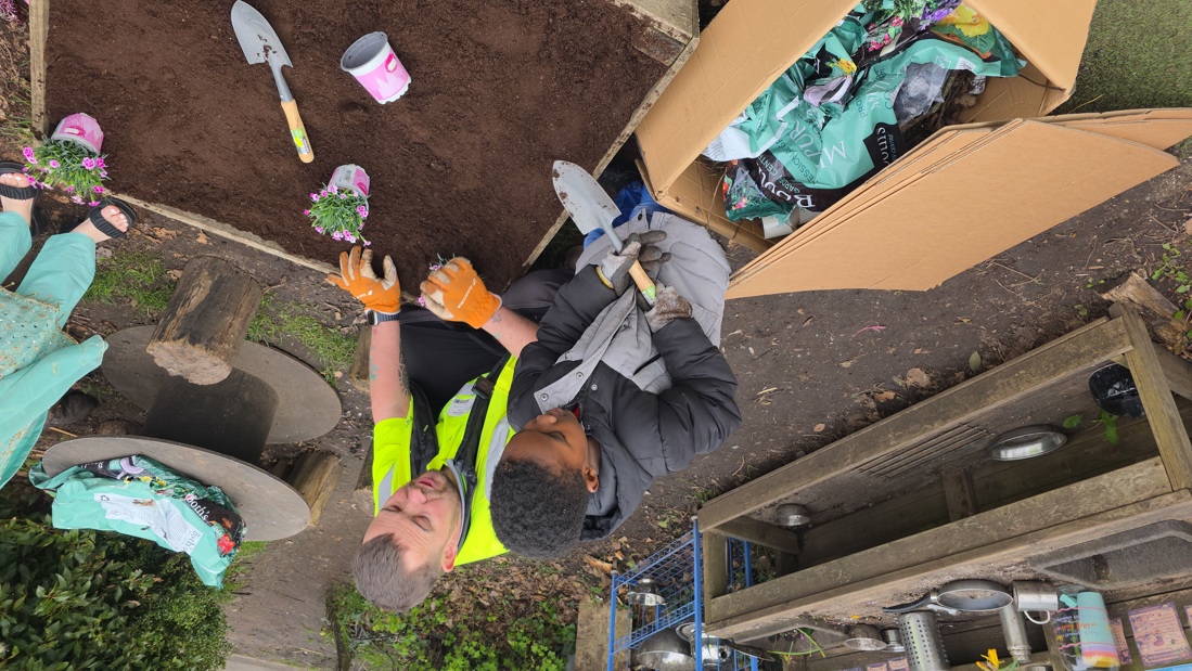 Adam Graham Guiding A Pupil With A Trowel Ahead Of Planting The Flowers